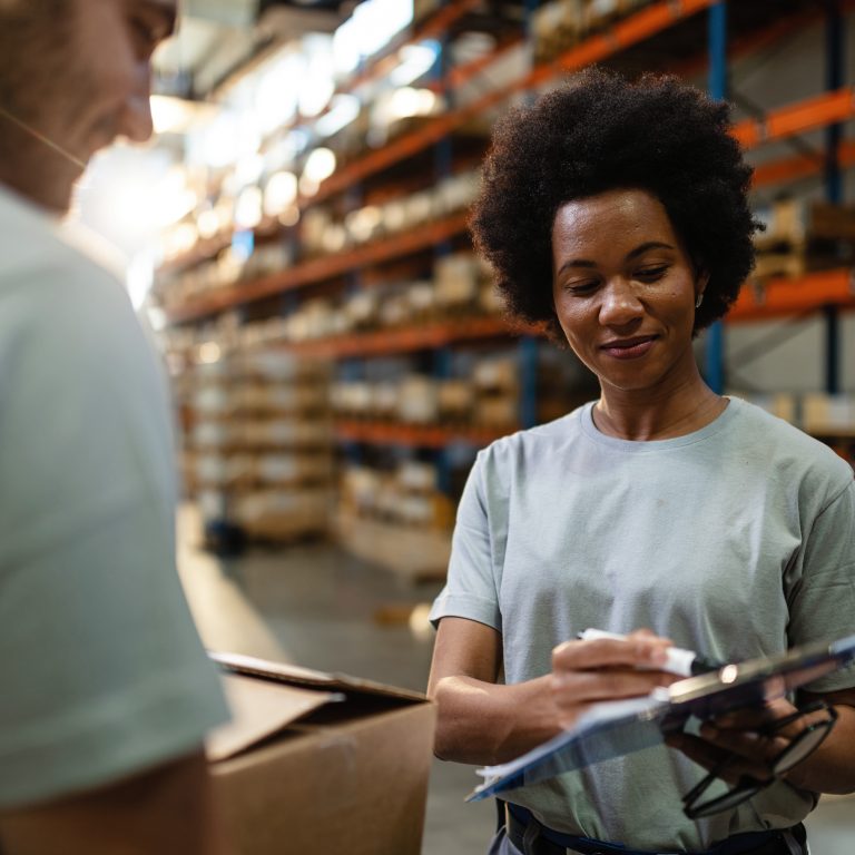 African American warehouse worker showing to her coworker check list of products for the shipment while working in distribution warehouse.